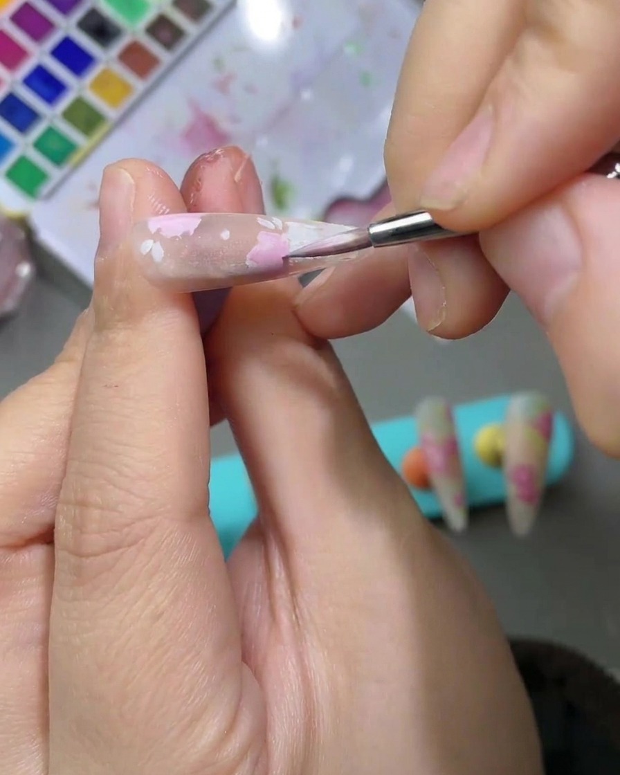 A detailed view of a nail art design featuring soft, light green watercolor leaves being painted next to pink rose petals.