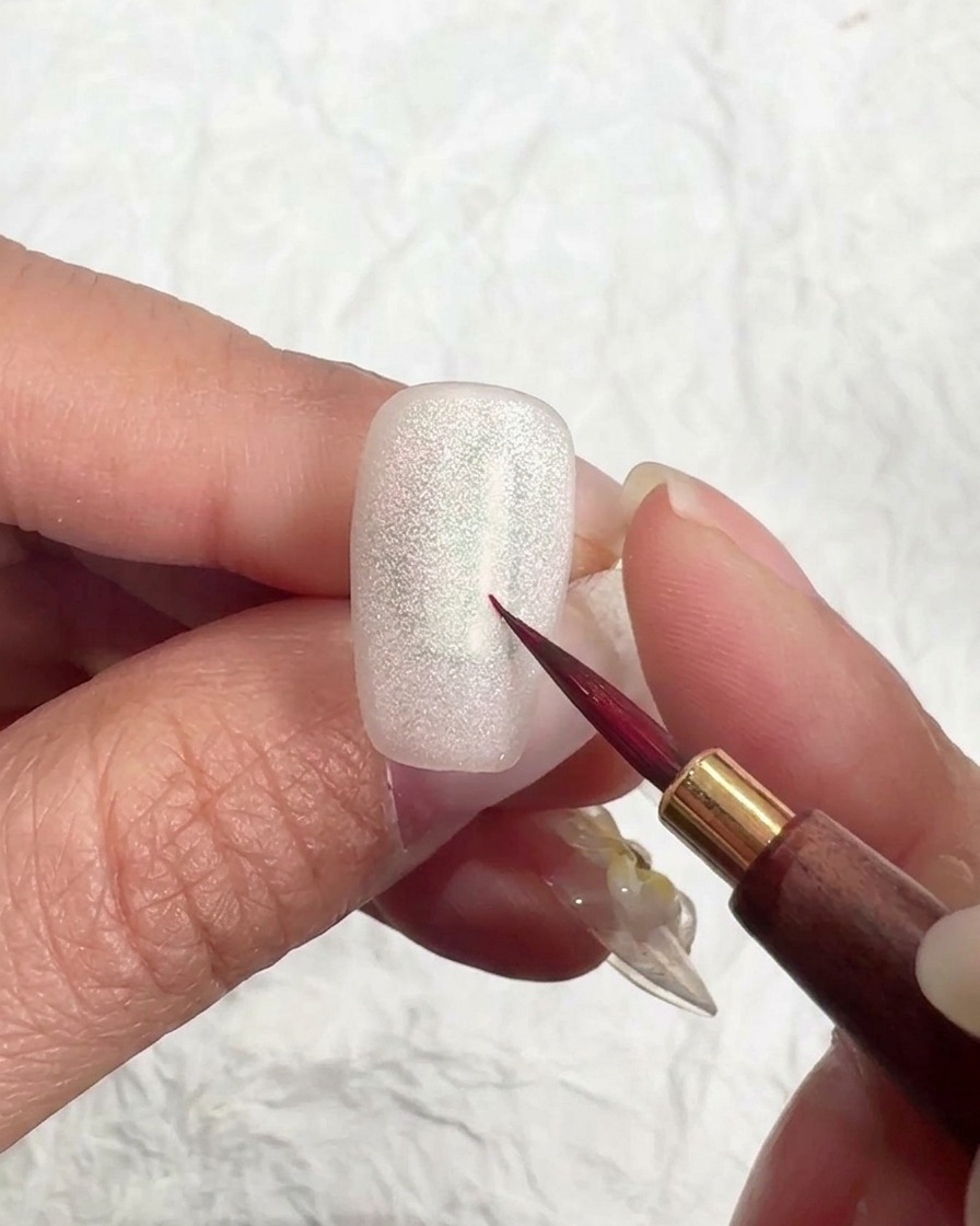 A nail brush applying a smooth coat of matte top gel over a shimmering cat-eye nail.