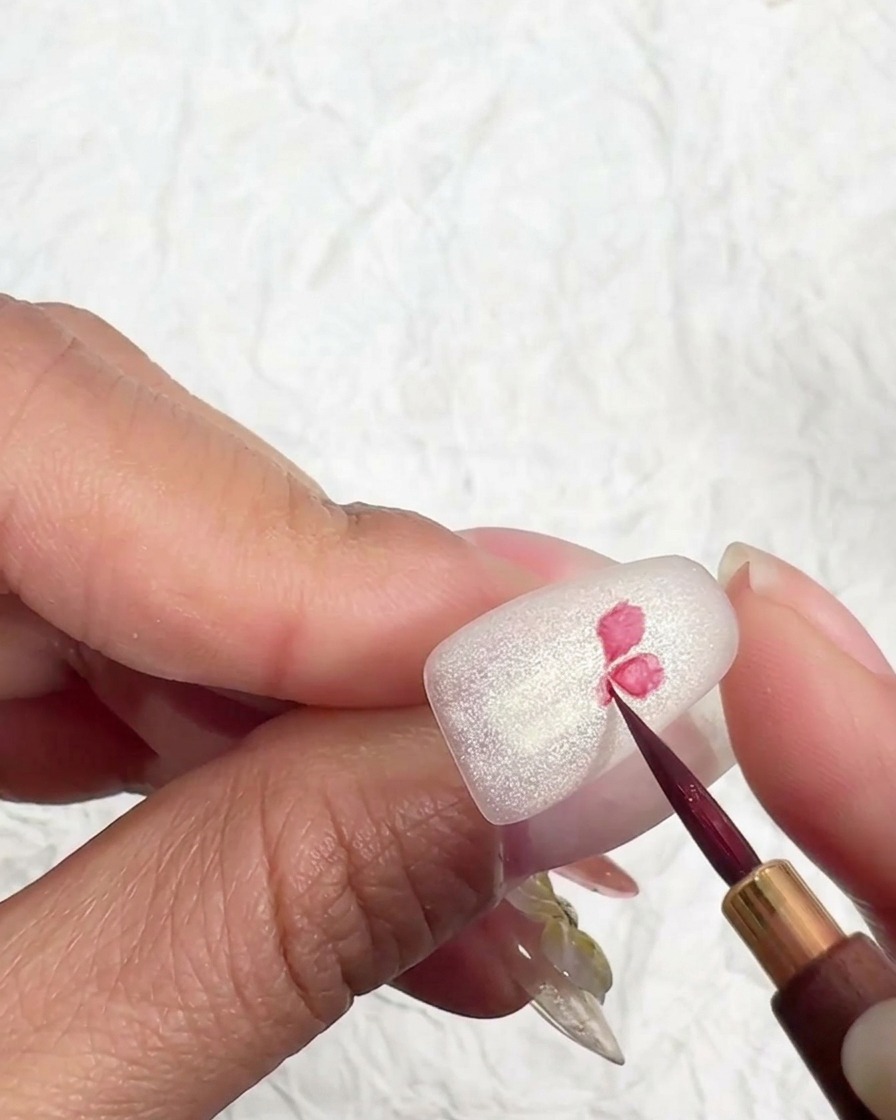 A fine nail art brush drawing the center of a pink cherry blossom on a matte nail surface.