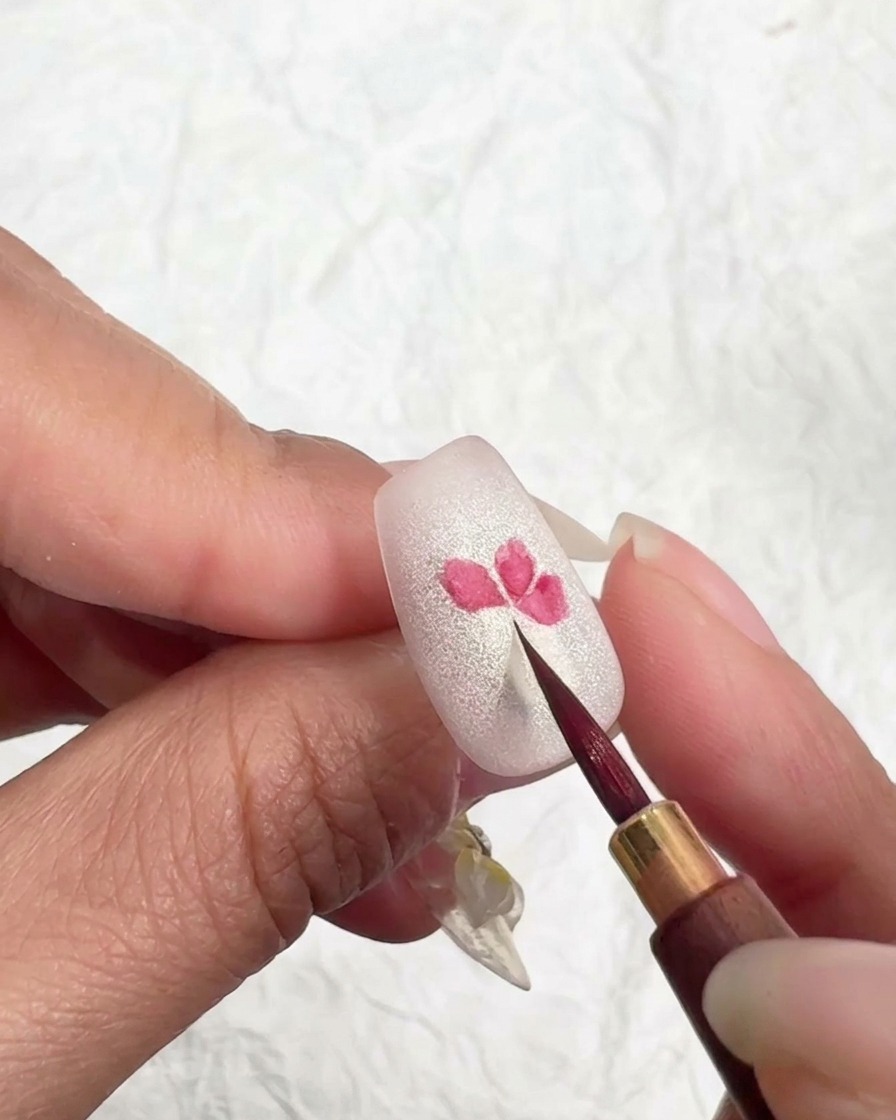 A fine brush lightly touching the edges of a pink flower petal on a nail to soften and blend the color.