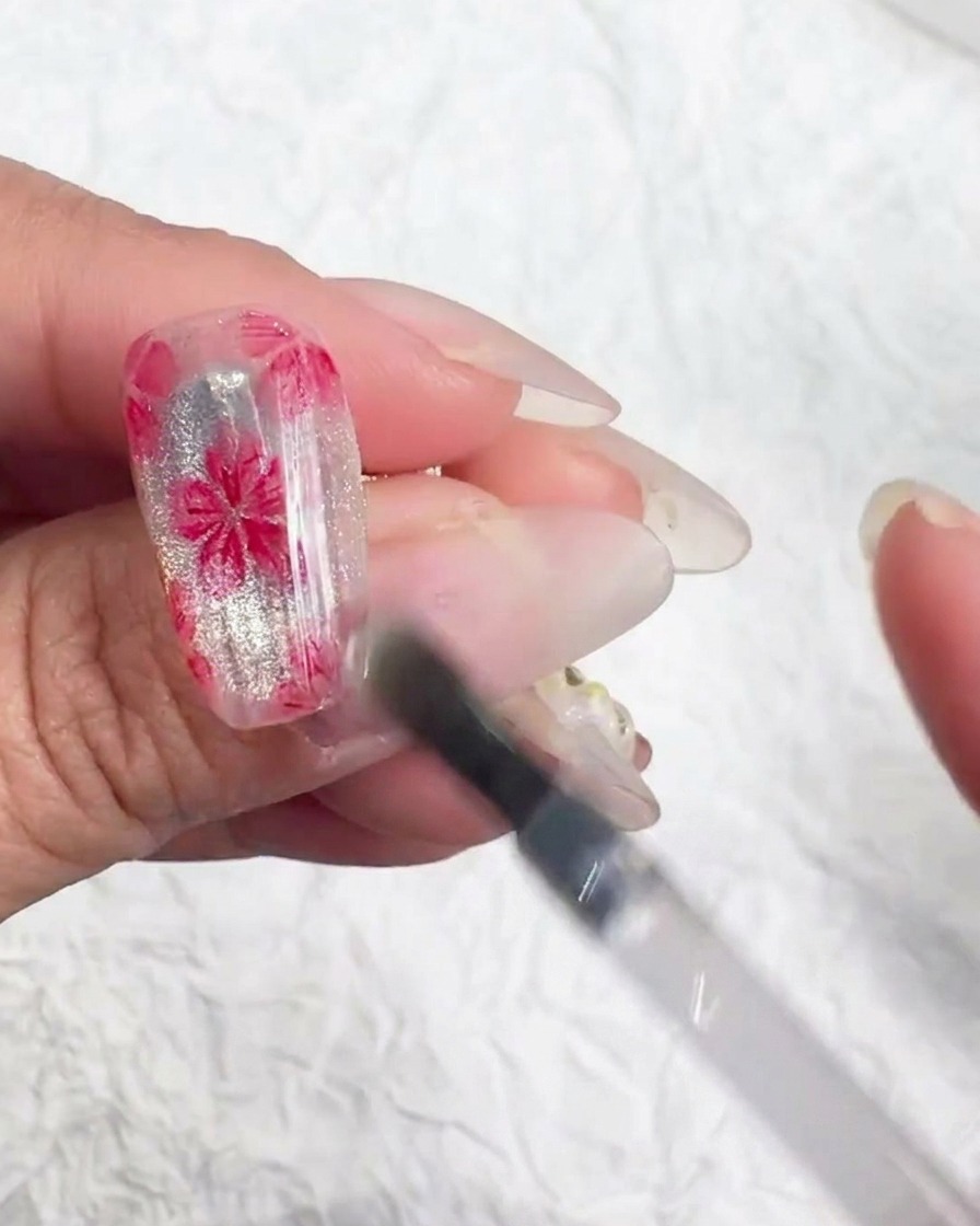 A small nail art tool applying tiny iridescent flakes to the center of a painted cherry blossom.