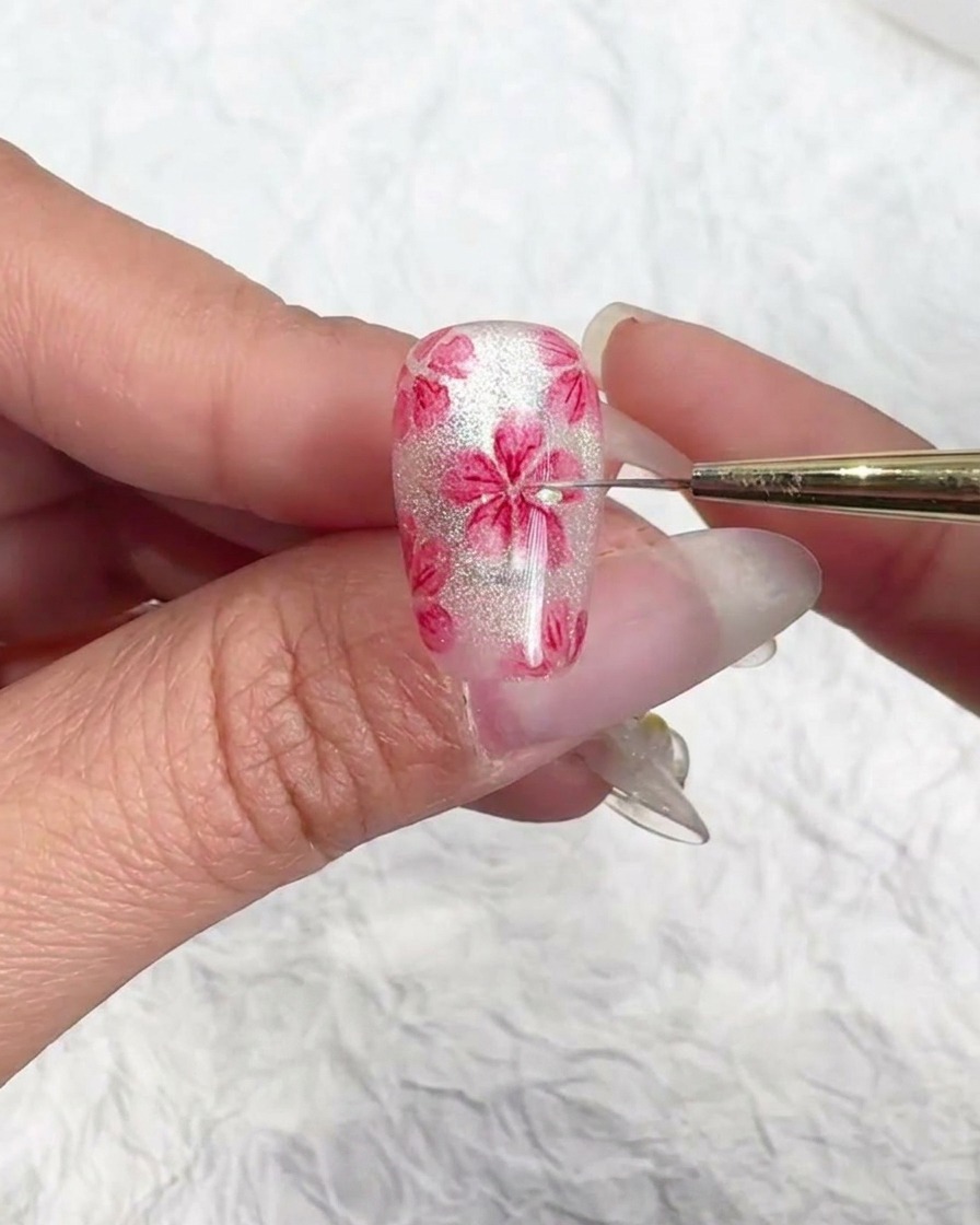 A close-up of a fine nail art brush adding tiny shiny flakes to the center of a hand-painted pink cherry blossom on a shimmering translucent cat-eye nail.