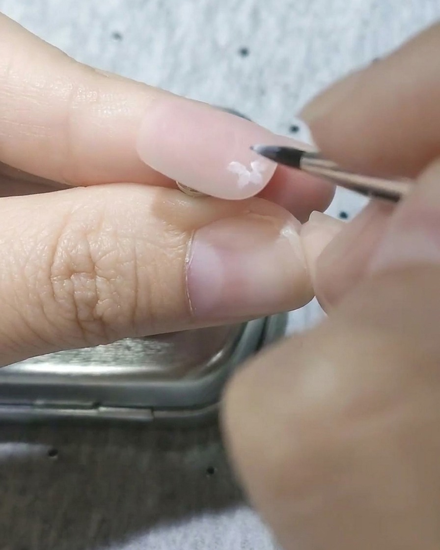 A close-up of a fine detail brush painting semi-transparent white cherry blossom petals onto a matte pink nail base.