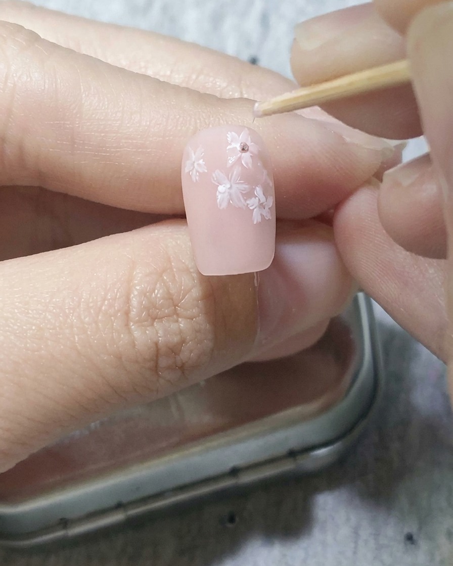 A toothpick gently placing dark pink dots into the center of white painted cherry blossoms on a soft pink nail.
