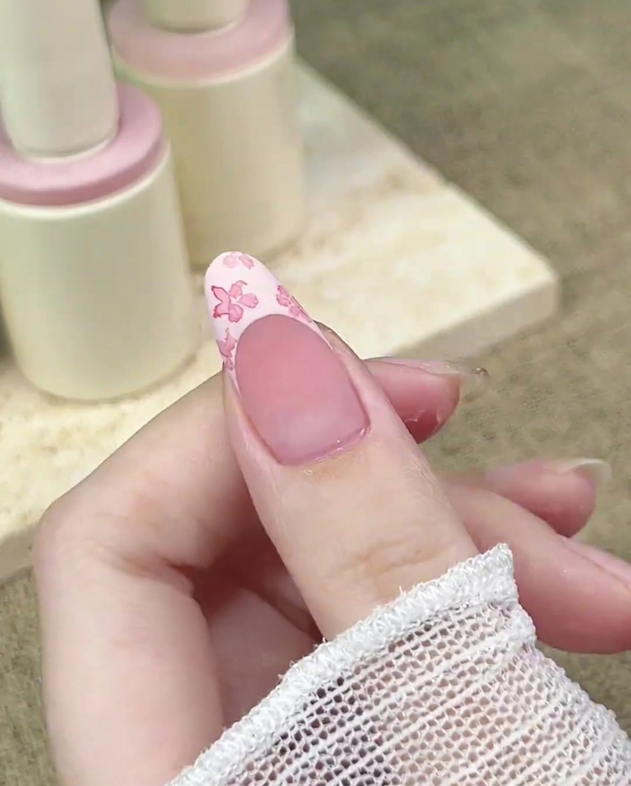 A close-up of a detail brush adding a concentrated dot of darker pink to the center of a floral design on a nail.