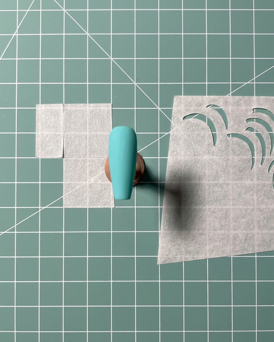 An adhesive stencil being applied to the center of a turquoise-painted nail tip.