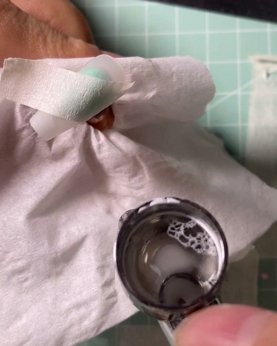 An airbrush tool spraying white pigment onto a nail tip held over a protective tissue.