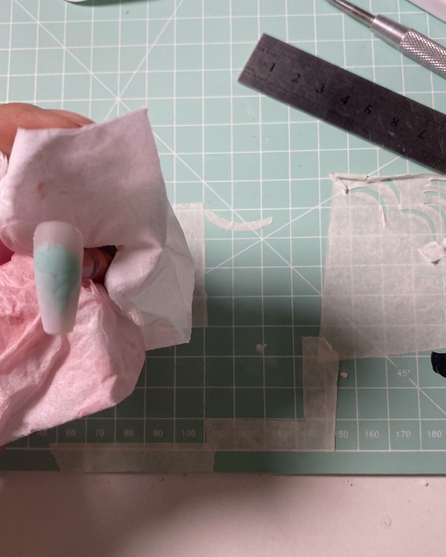 An airbrush tool being used to apply a soft dusty rose color onto a nail tip.
