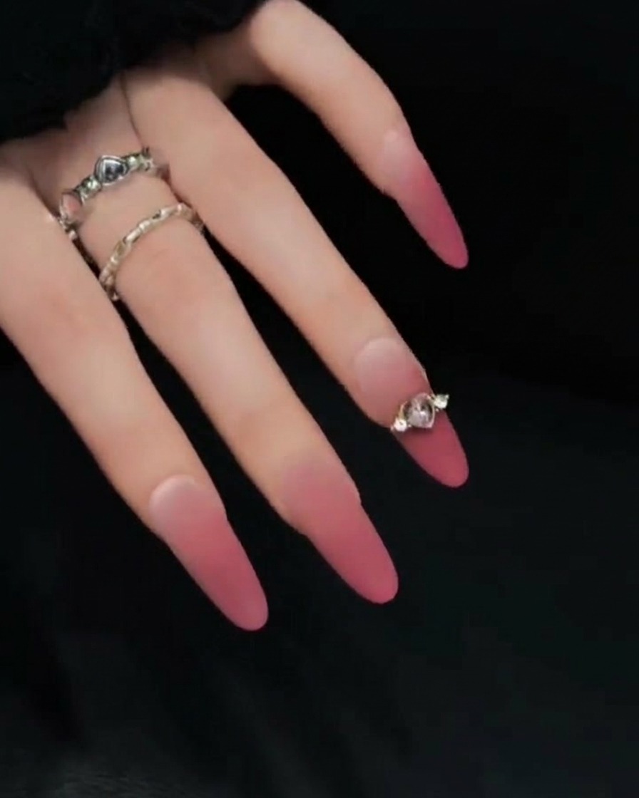 A close-up of a hand posed against a dark background, showing the side profile of long pink and nude gradient press-on nails to highlight their length and curvature.