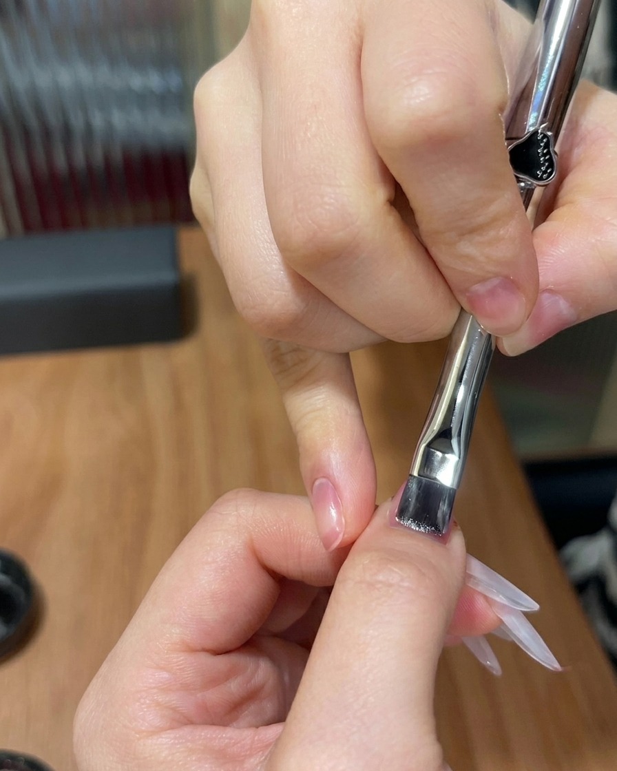 A flat brush applying a thin layer of shimmering cat-eye gel over a nail with a pre-existing pink gradient.