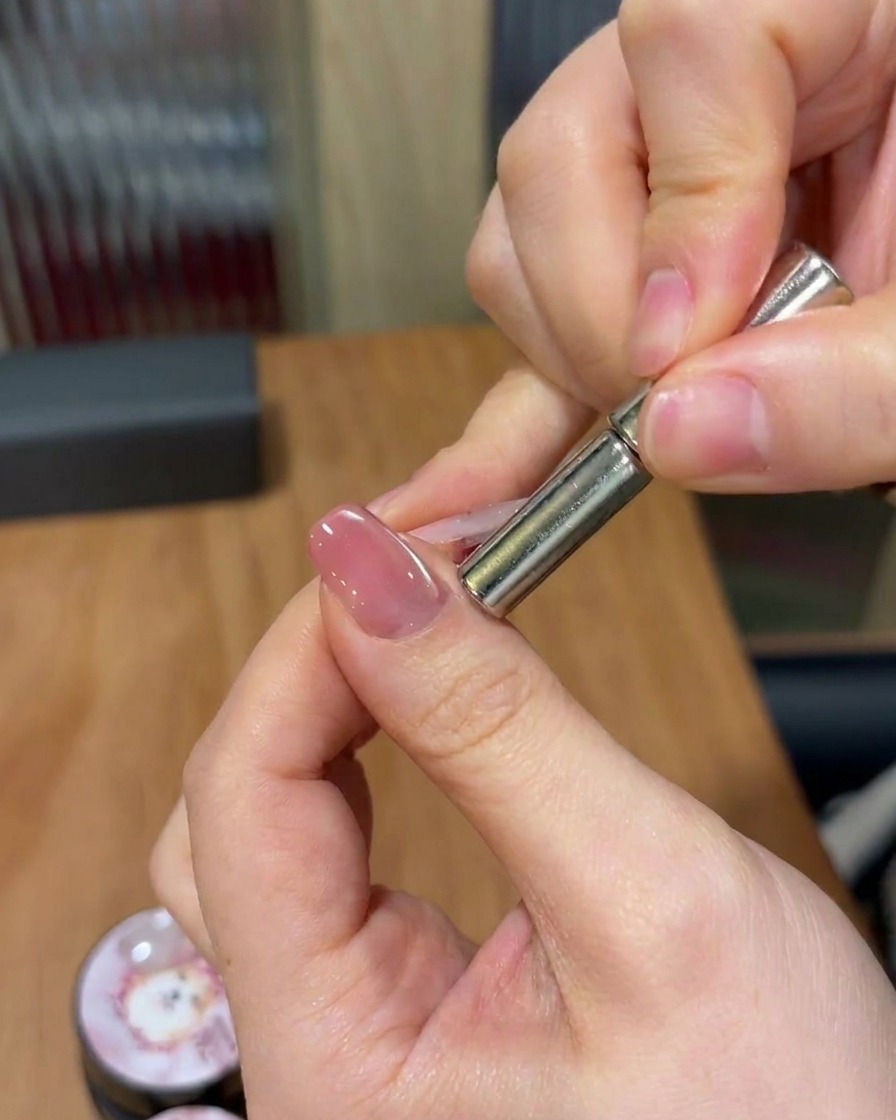 A hand holding a cylindrical magnet above a nail, sweeping it to manipulate magnetic particles into a shimmering diagonal band.