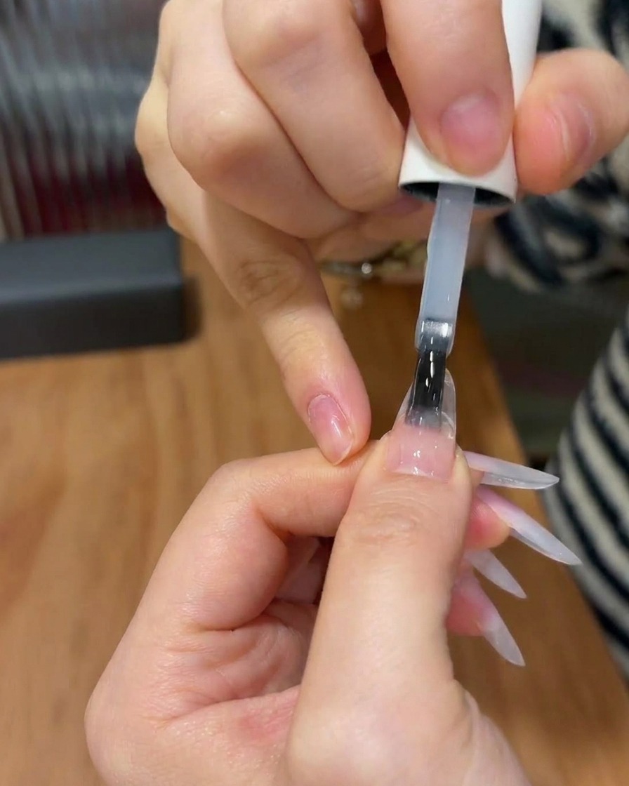 A nail brush applying a clear, glossy top coat to a nail with a finished pink gradient design.