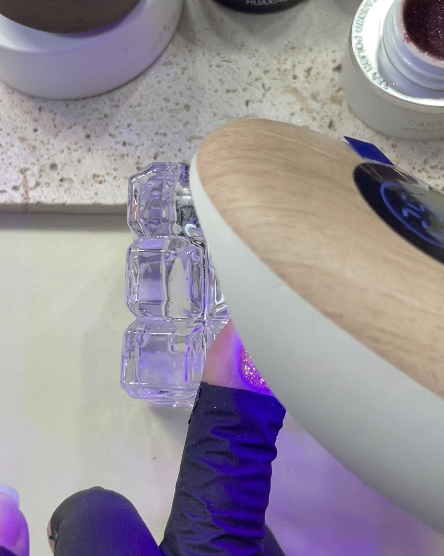A hand positioned under a UV/LED lamp while curing a nail with pink glitter applied.