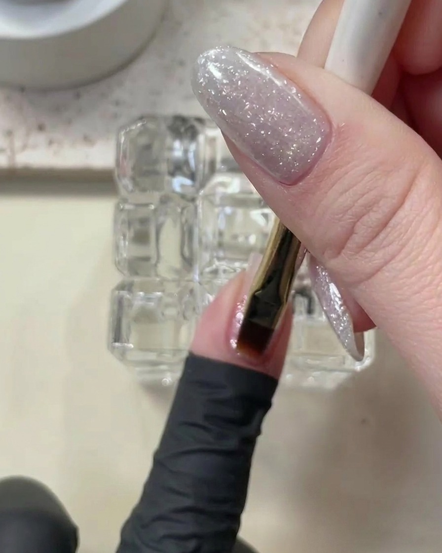 A brush applying a thin, clear layer of builder gel onto a glitter-coated nail.