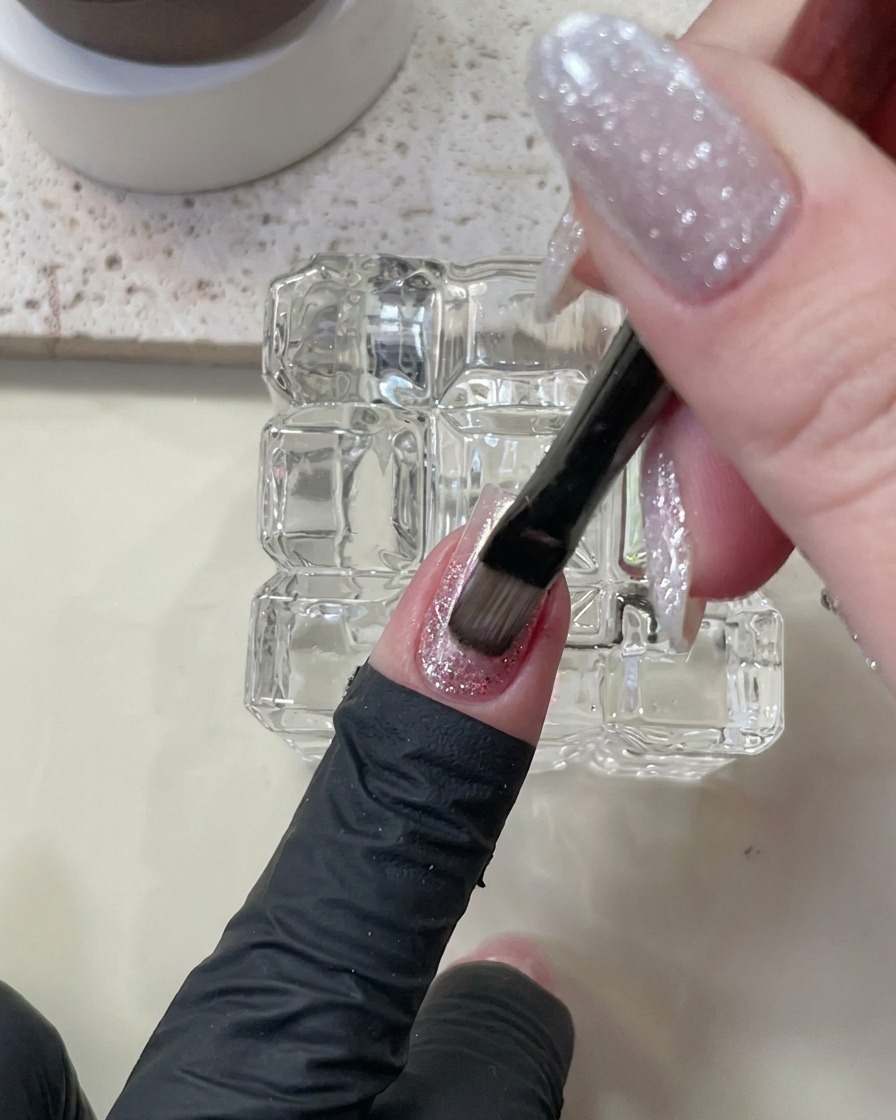 A brush applying nude cat-eye gel polish over the existing nail layers.