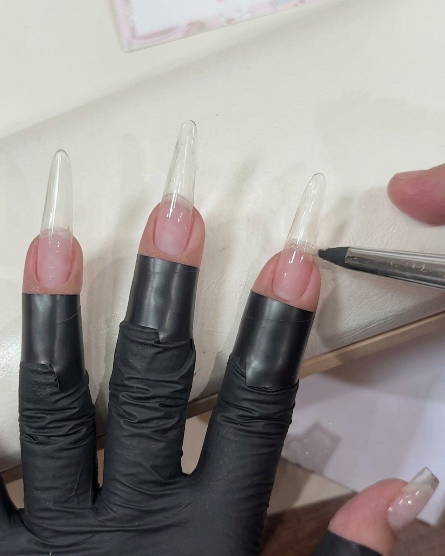 Applying a sheer light pink gel to a long clear nail extension using a flat brush.