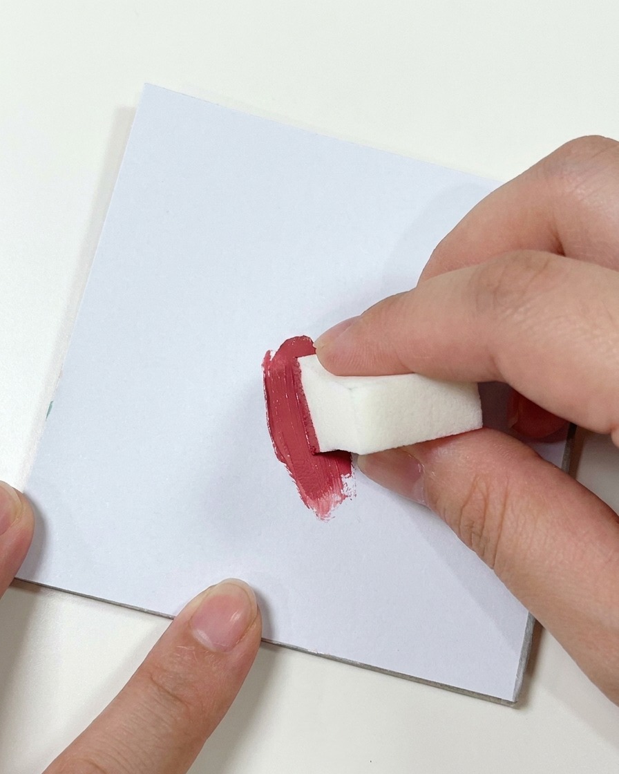 A hand pressing a small white makeup sponge into a smudge of dark pink gel on a flat white palette surface.