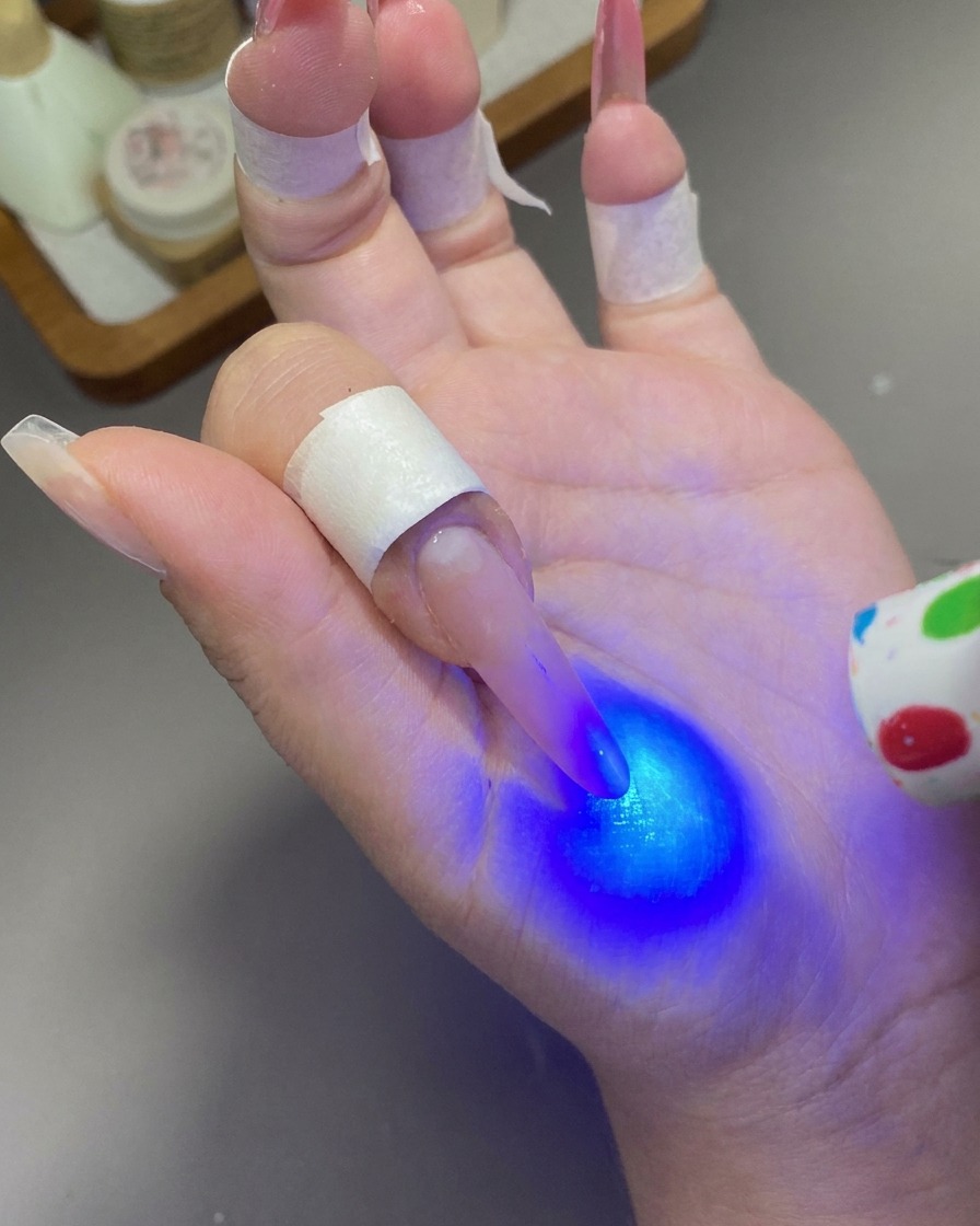 A mini handheld UV/LED lamp shining blue light to cure the gel on a fingernail.