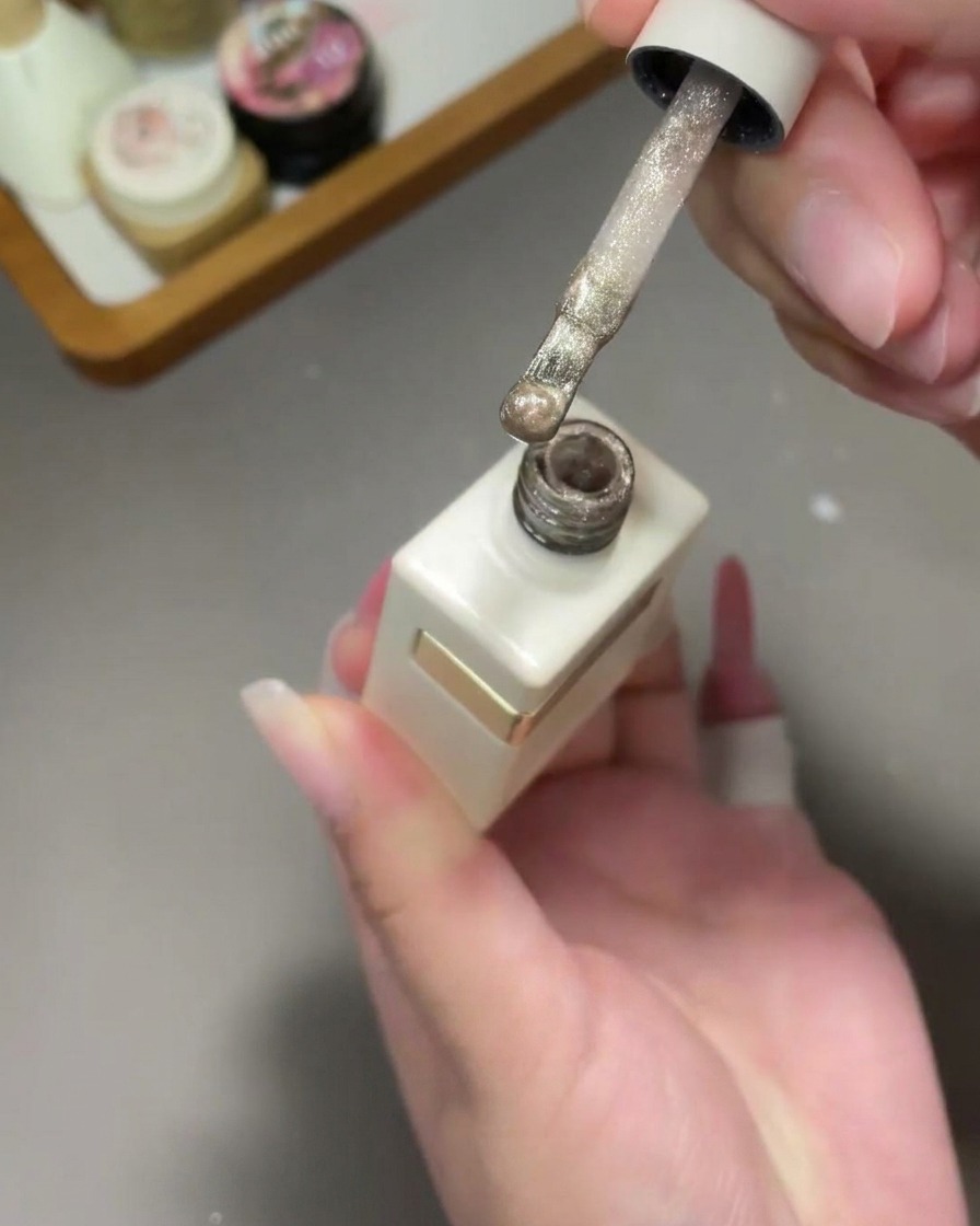 A brush applying a shimmering gold cat-eye gel polish over a pink gradient nail.