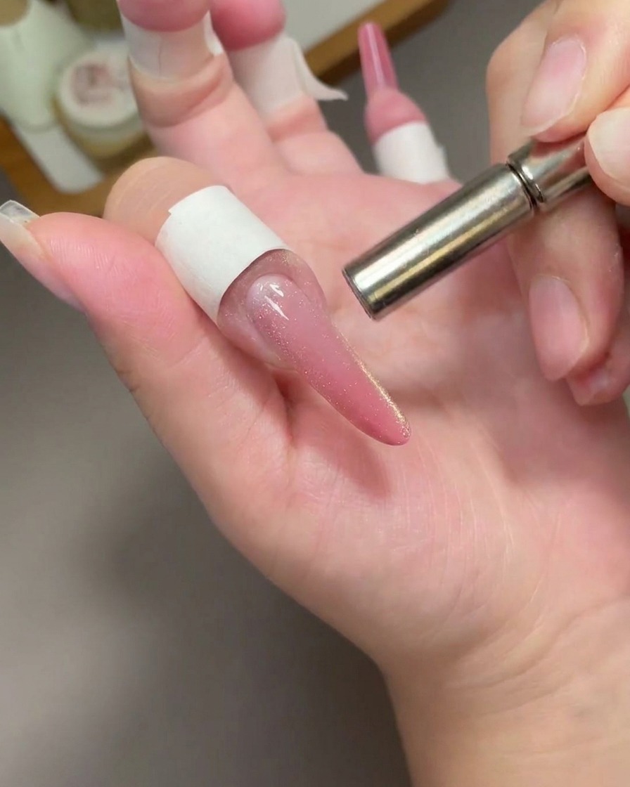 A close-up view showing a cylindrical metal magnet hovering over a long, pink cat-eye gel nail to move the magnetic pigment.