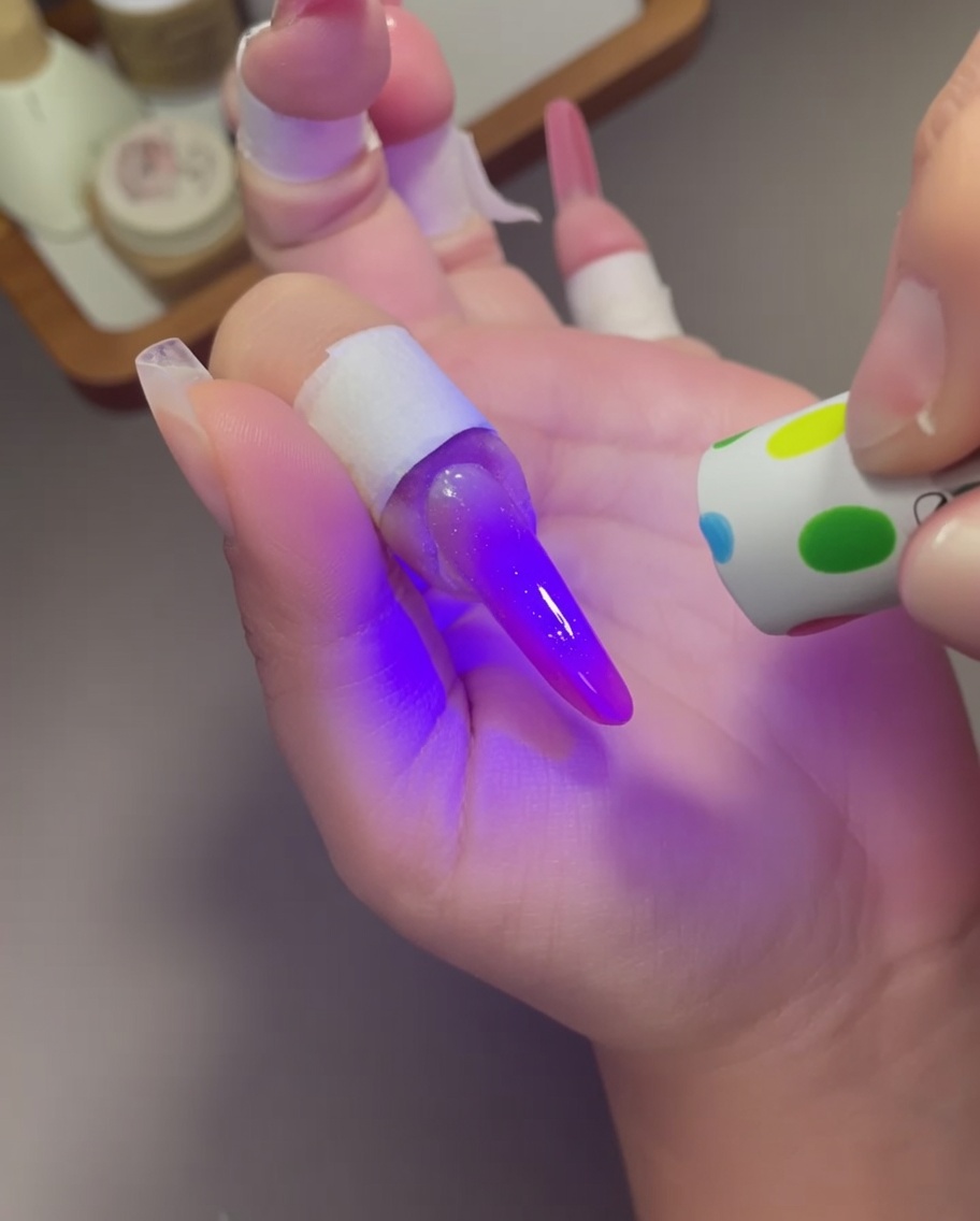 A small, polka-dot patterned handheld UV lamp held over a finger with a bright purple-pink cat-eye nail design.