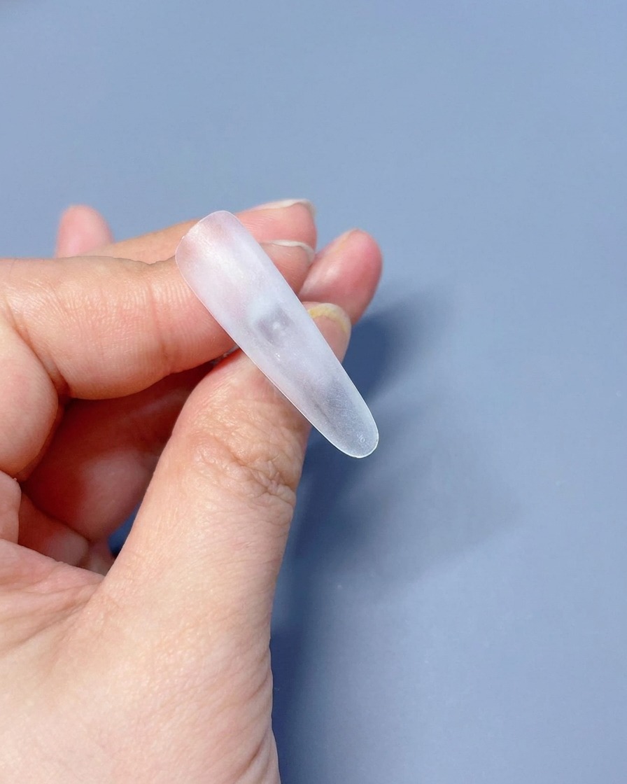 A close-up of a clear, unpainted press-on nail tip being held between fingers, ready for the base coat application.