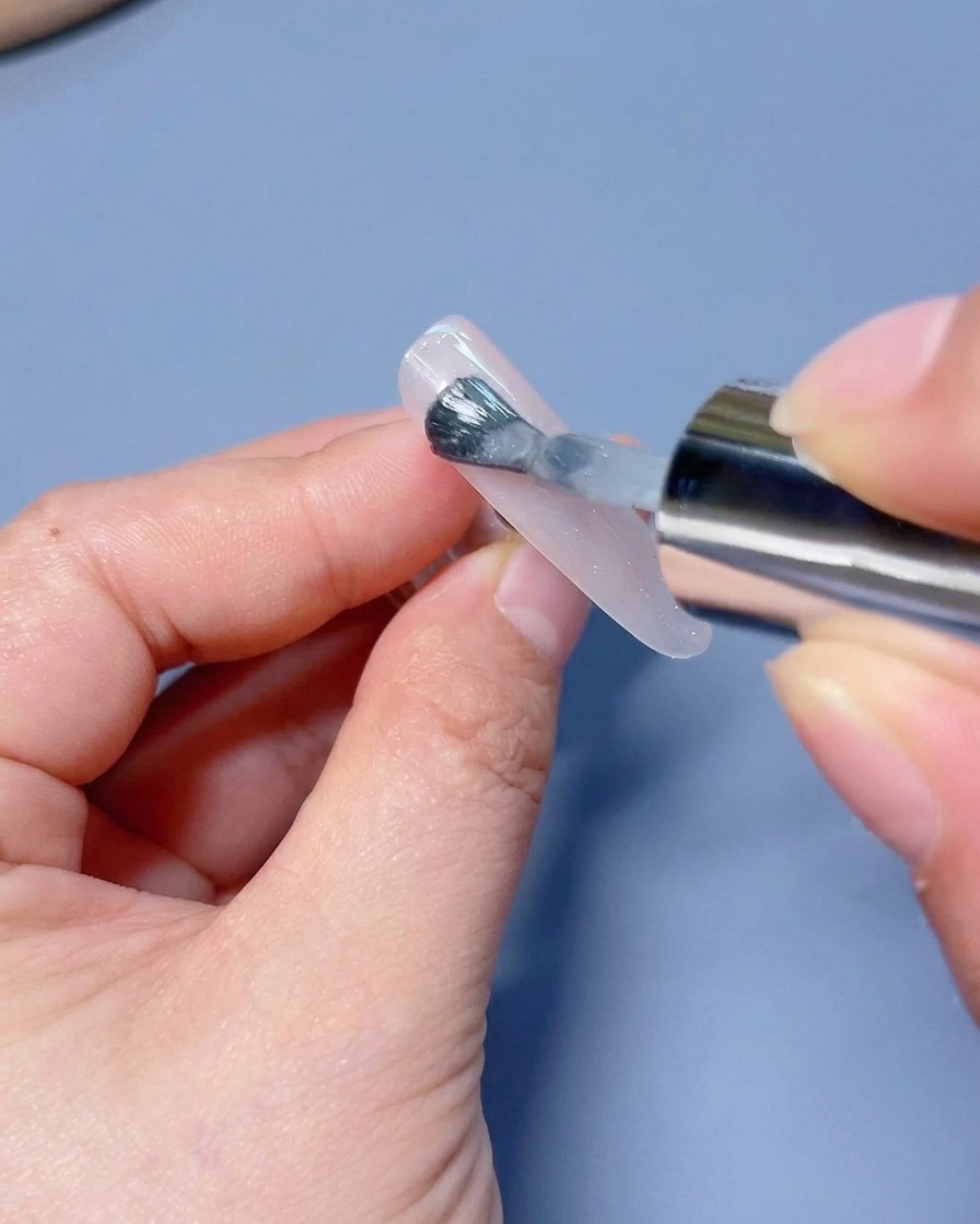 A brush applying a coat of shimmering white cat-eye gel polish to a press-on nail tip.