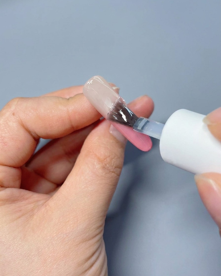 A brush applying a clear, glossy top coat over a soft pink gradient nail surface.