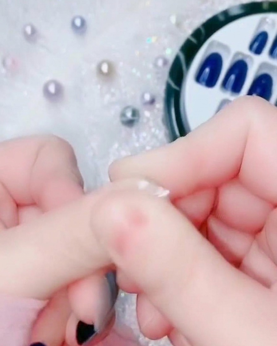 A close-up of a finger pressing the base of an electric blue press-on nail against the natural cuticle area.