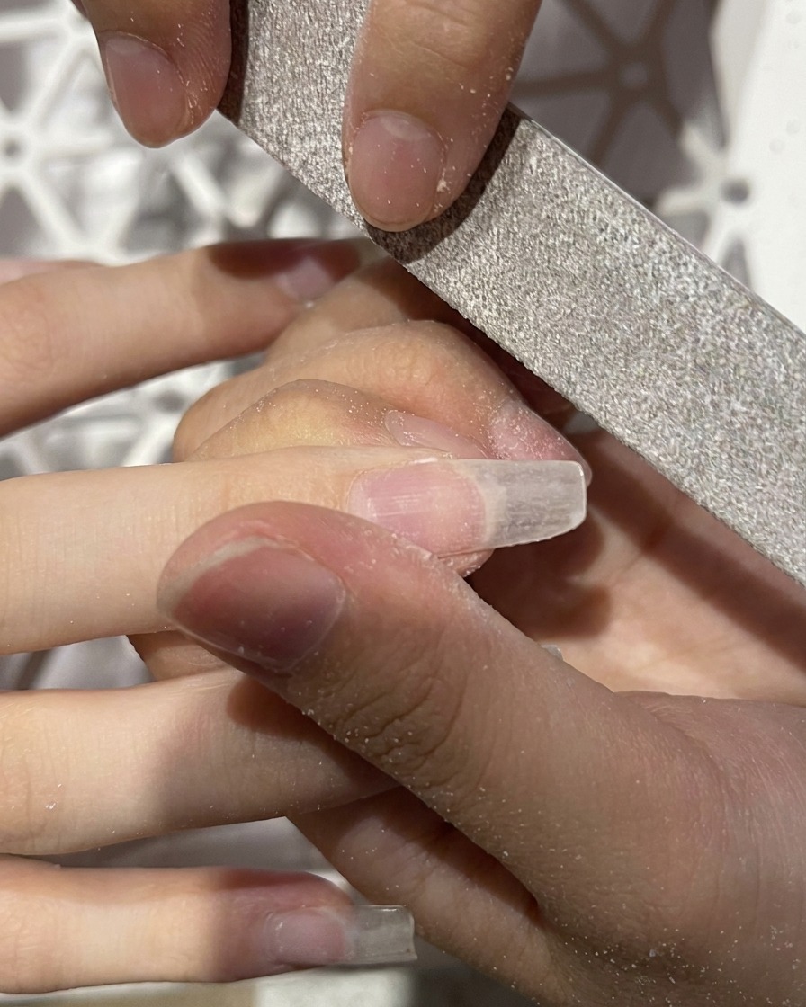 A close-up of a hand file being used to shape the free edge of an artificial nail extension into an almond shape.