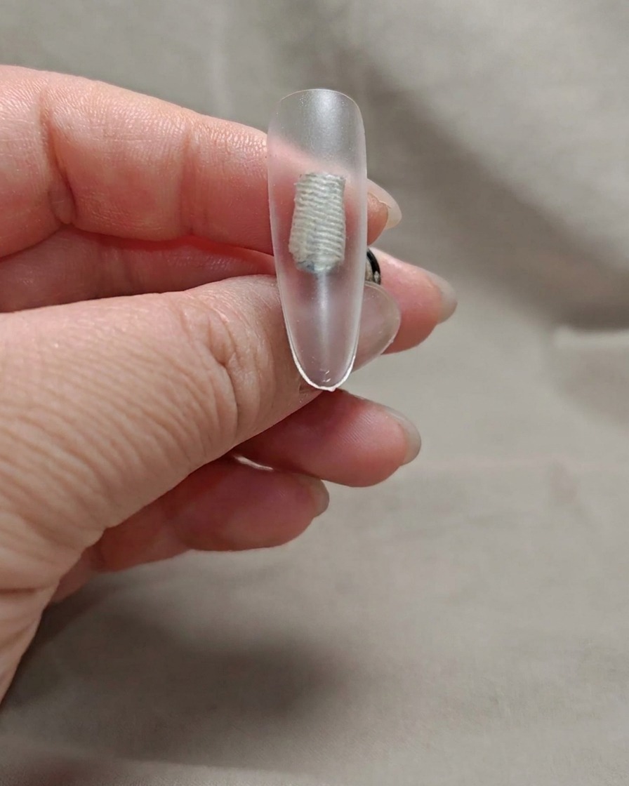A brush applying a thin layer of sheer nude pink gel to a nail tip.