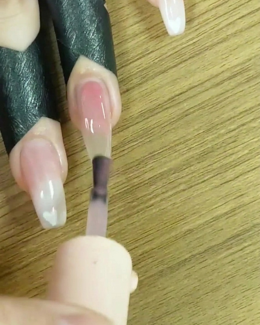 A brush applying sheer nude gel polish to the full nail surface.