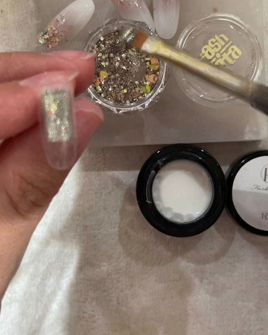 A brush applying chunky silver and iridescent glitter to the top half of a clear nail tip.