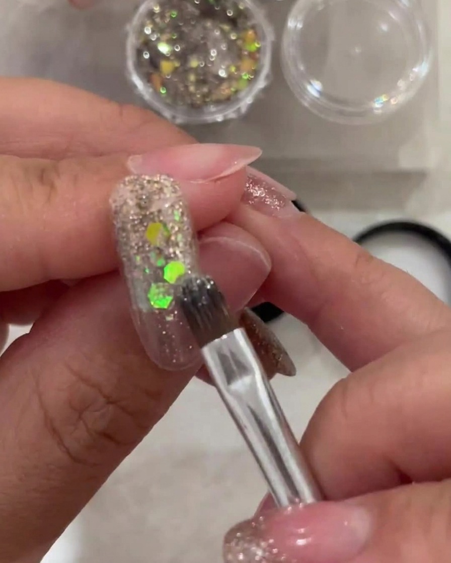 A brush applying a thin layer of sheer white gel to the tip of a nail, blending toward the glitter-covered cuticle area.