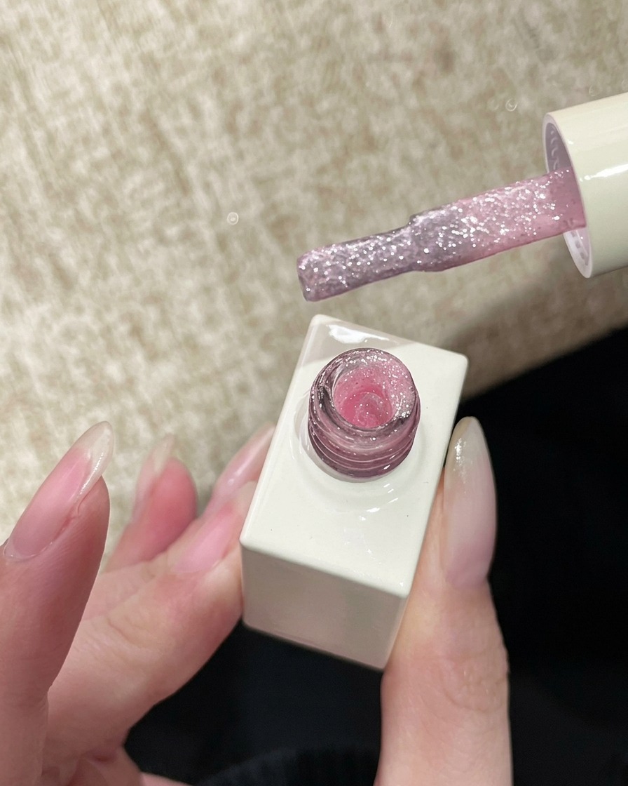 A nail polish brush holding a sheer, sparkling pink glitter gel, ready to be applied to the nail.