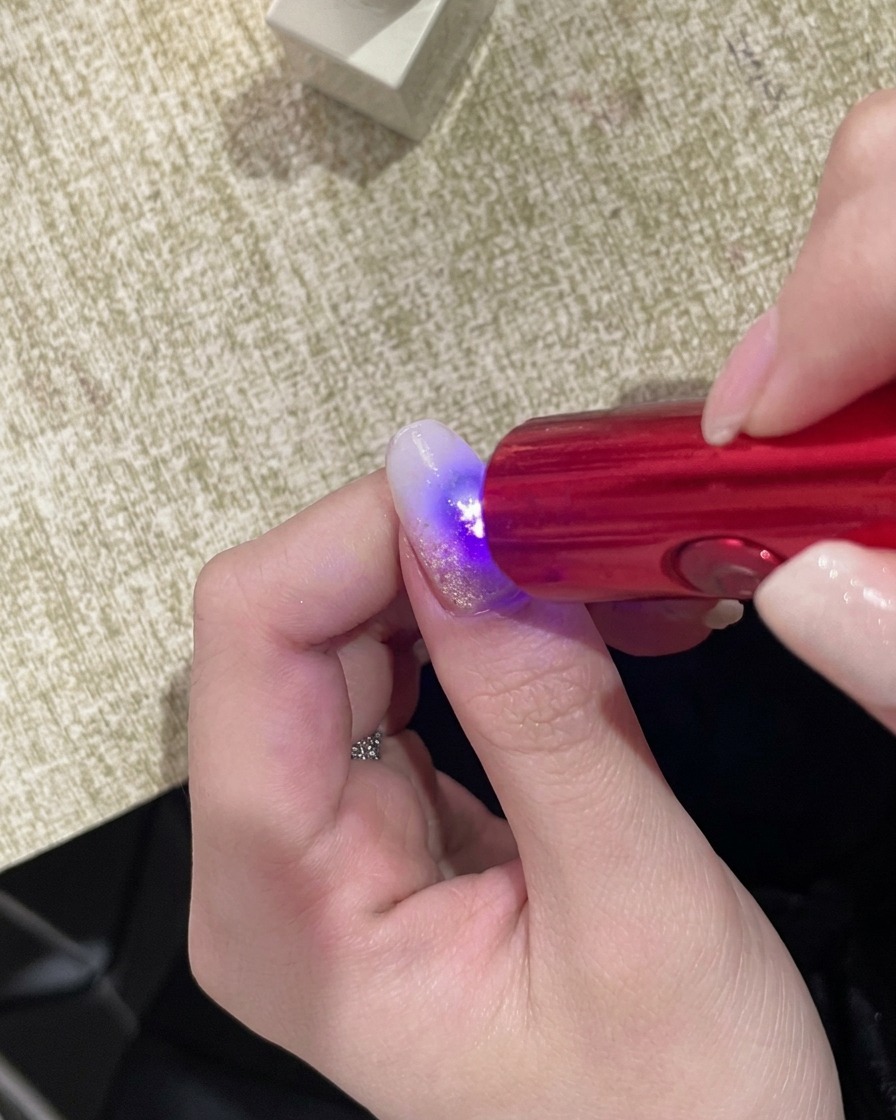 A hand positioned under a compact handheld UV lamp, with the nail surface glowing under the light.