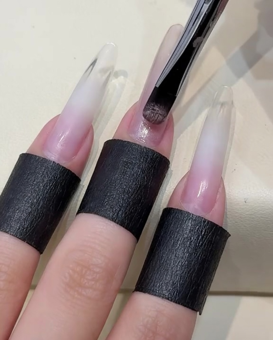 A brush applying a layer of light, nude-toned cat-eye gel polish to the nail.