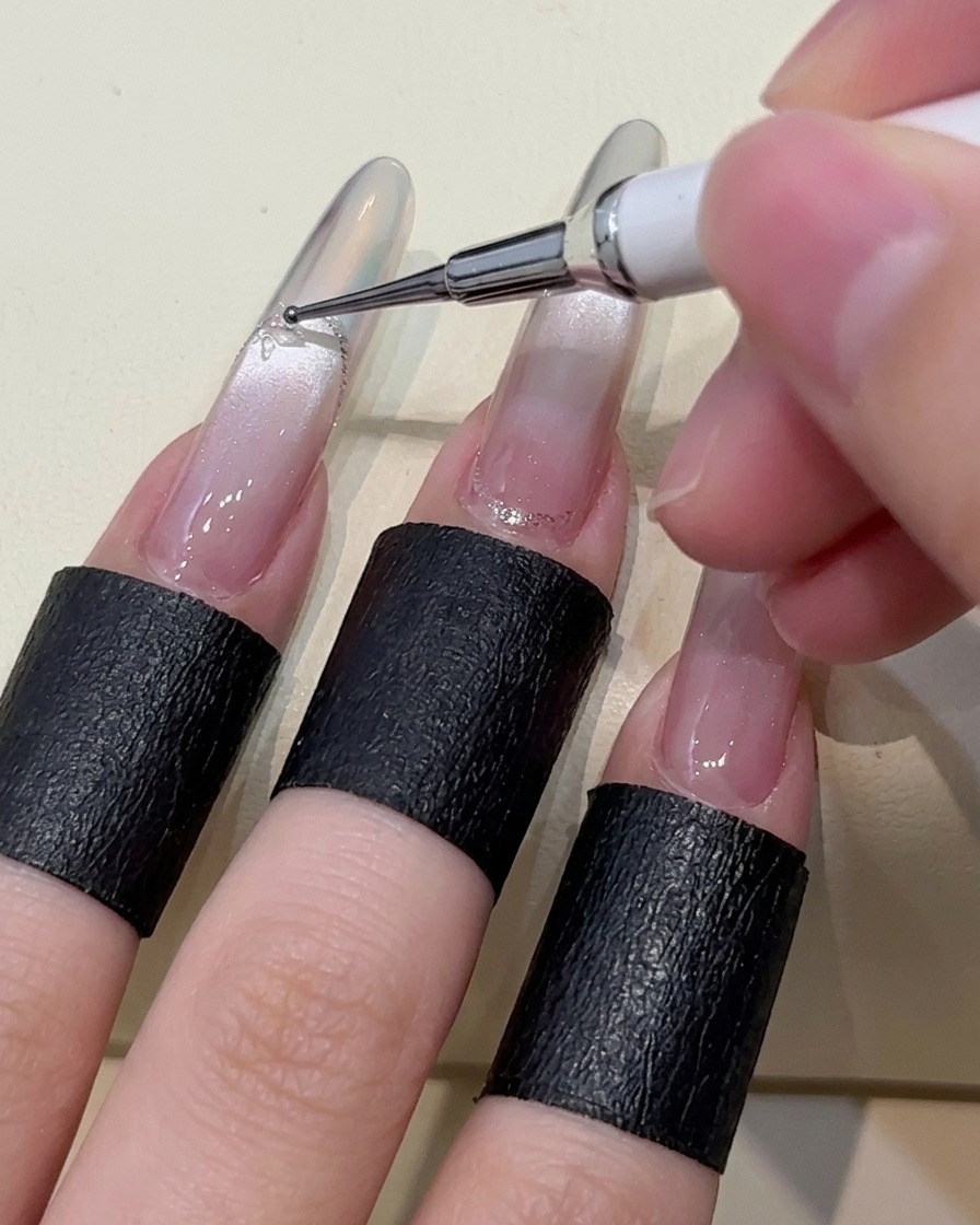 A close-up of a dotting tool placing small beads of clear adhesive gel onto the surface of a manicured nail.