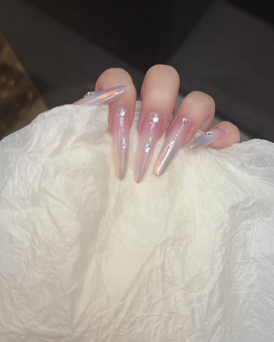 A close-up of a nail brush applying a nude base color coat to the nail plate.