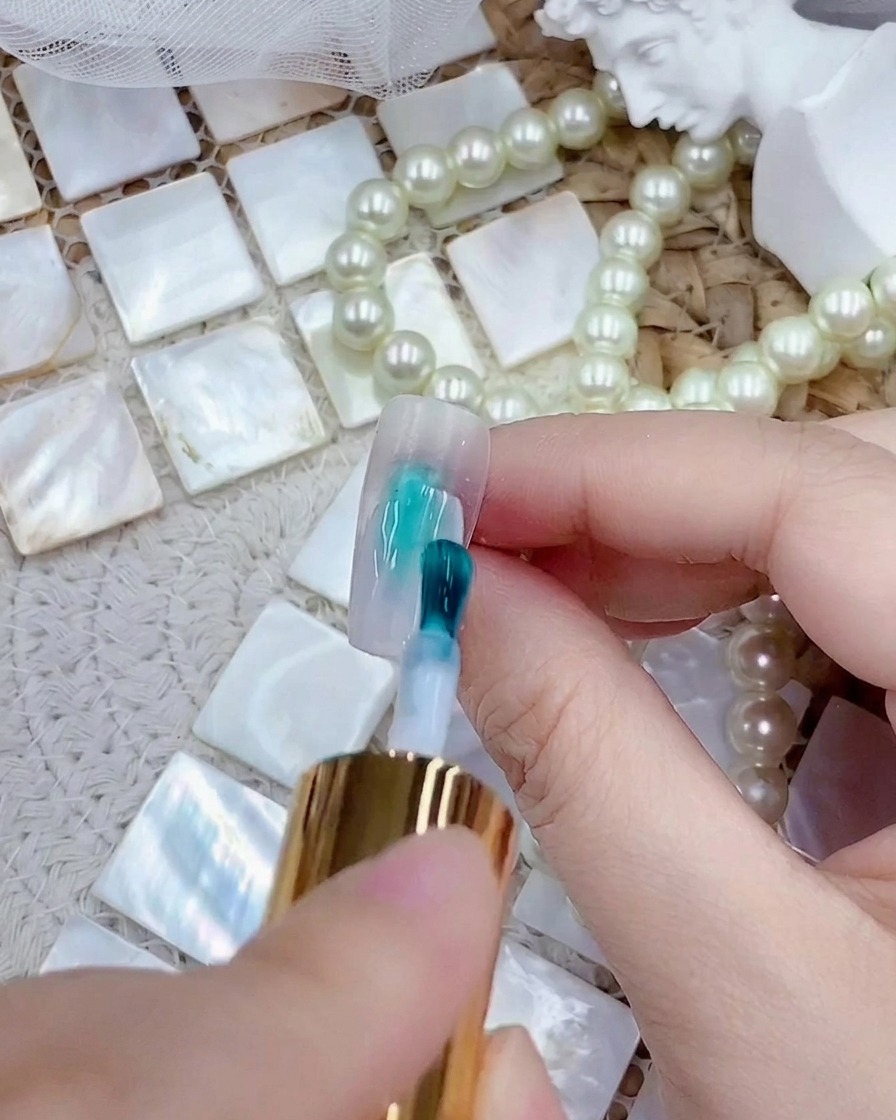 A brush applying a coat of milky white gel polish to a clear press-on nail tip.