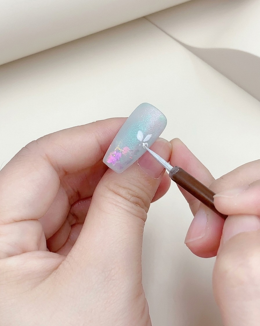A close-up of a fine brush adding a silver outline to a white painted flower on a nail.
