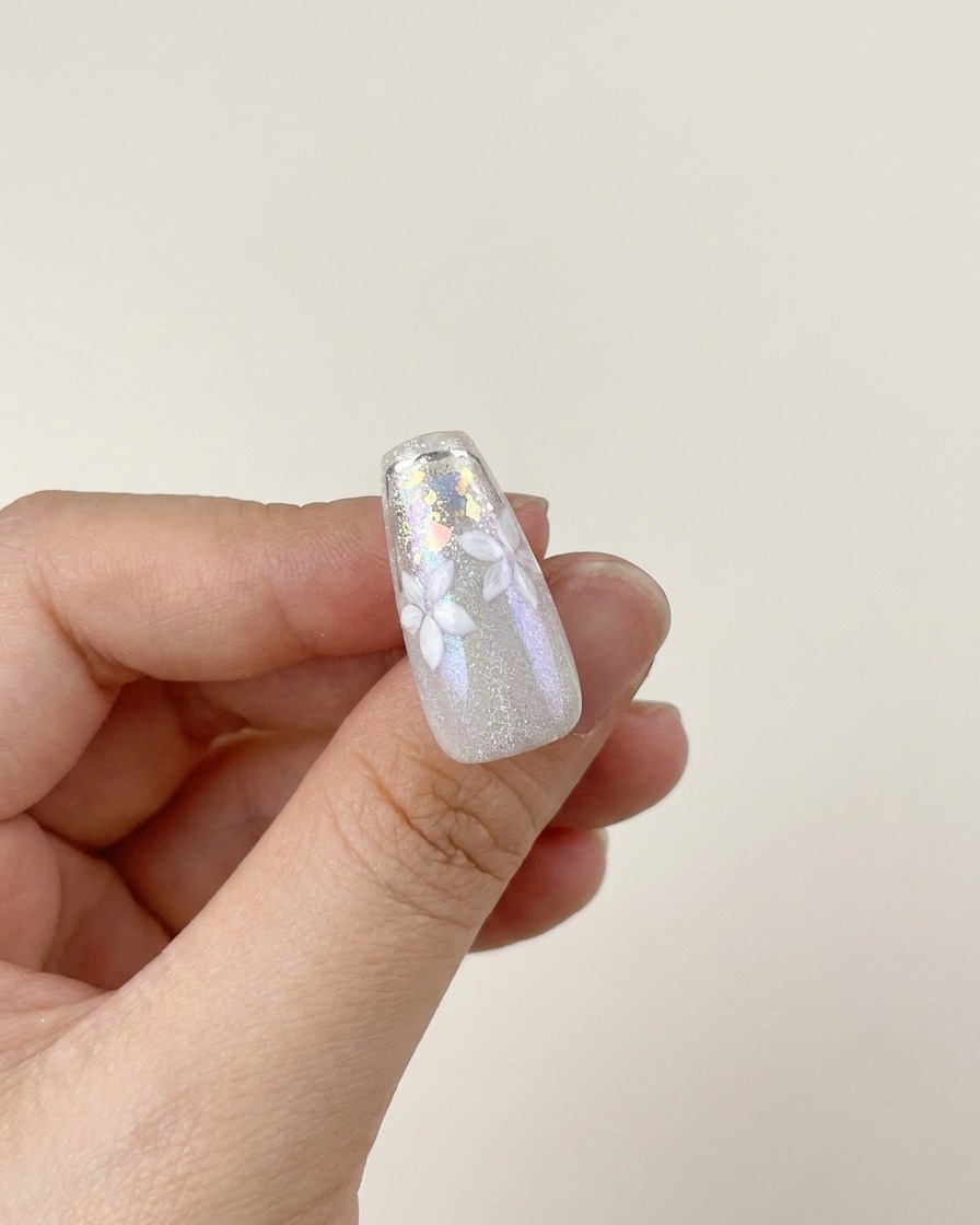 A brush applying a layer of clear, high-shine glossy top coat over a finished nail design featuring white flowers and silver flakes.