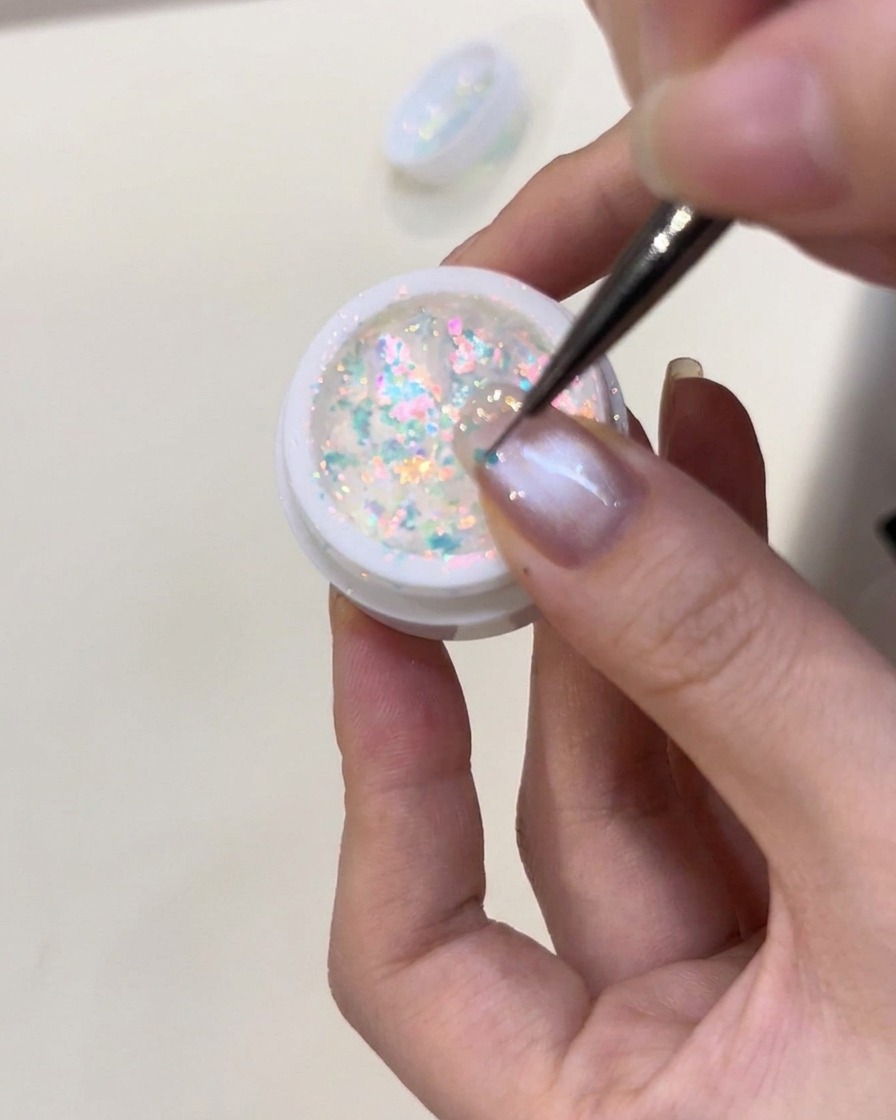 Tweezers carefully placing small, glowing blue iridescent flakes onto a nail coated in wet, clear builder gel.