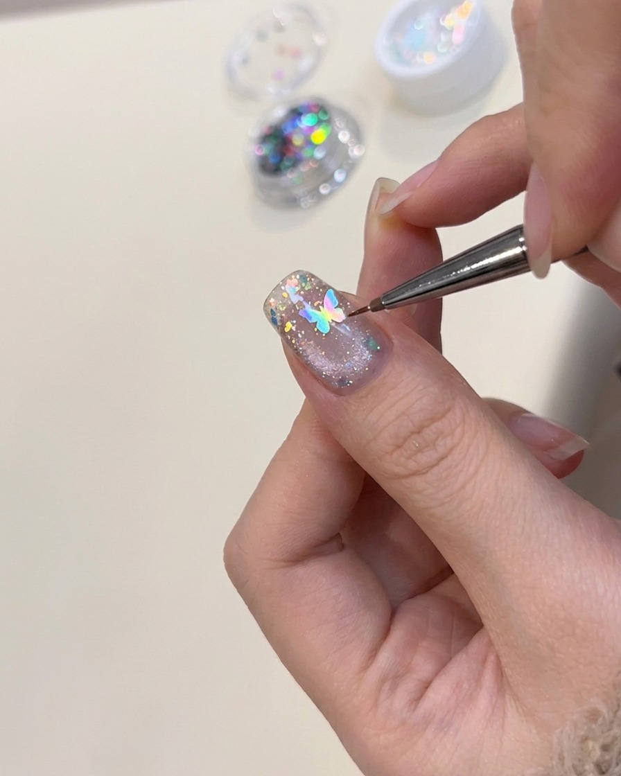 A pair of tweezers positioning a small, holographic butterfly-shaped nail charm onto a decorated nail.