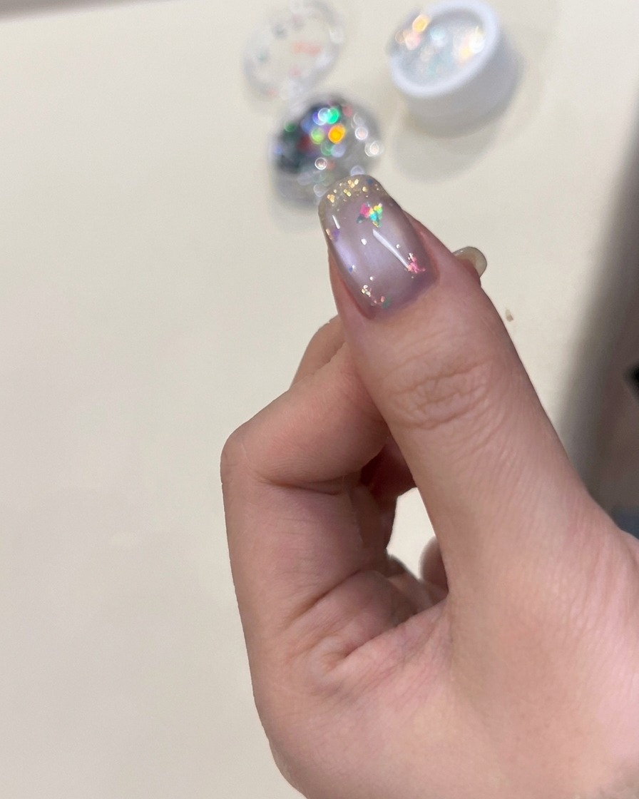 A nail brush applying a clear, glossy top coat over a nail decorated with iridescent flakes and a butterfly charm.