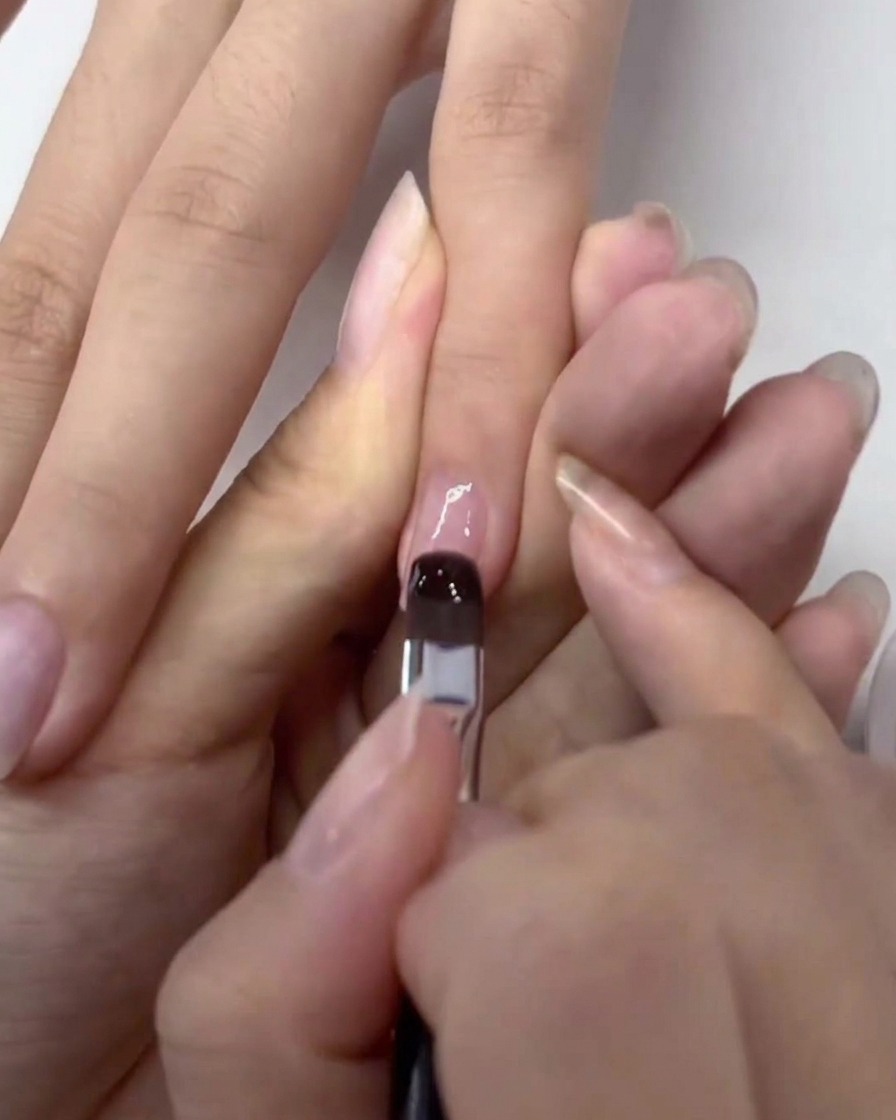 A brush applying a clear layer of gel base coat onto a natural, prepped fingernail.