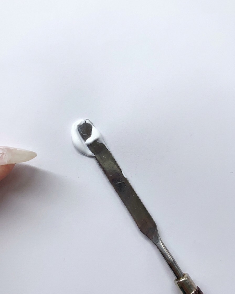 A metal spatula mixing a dollop of white gel and blue gel together on a clean white surface to create a light blue custom shade.