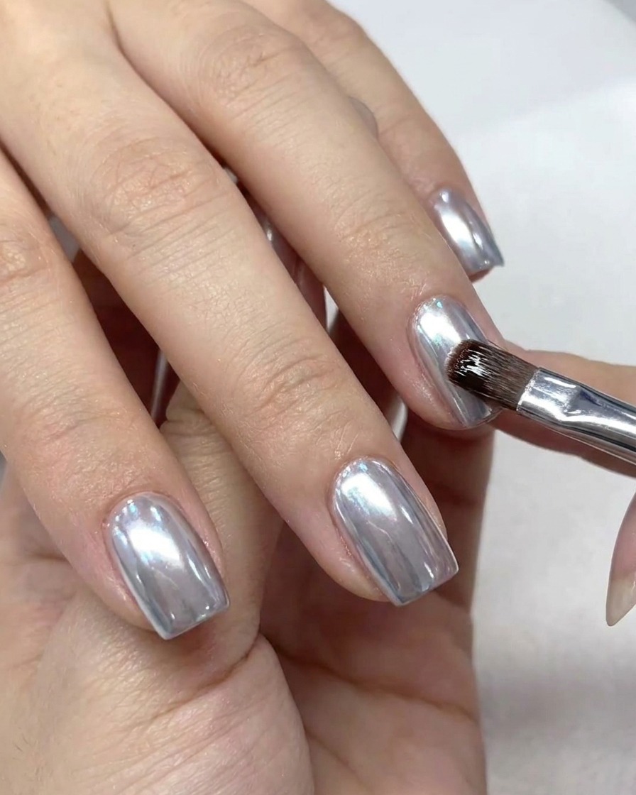A small makeup brush dusting excess chrome powder from the cuticle and skin around the nail.