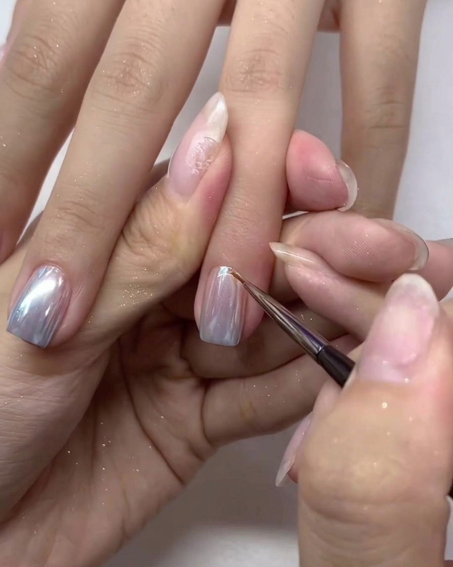 A small nail brush carefully applying a final layer of top coat over the silver chrome nails.