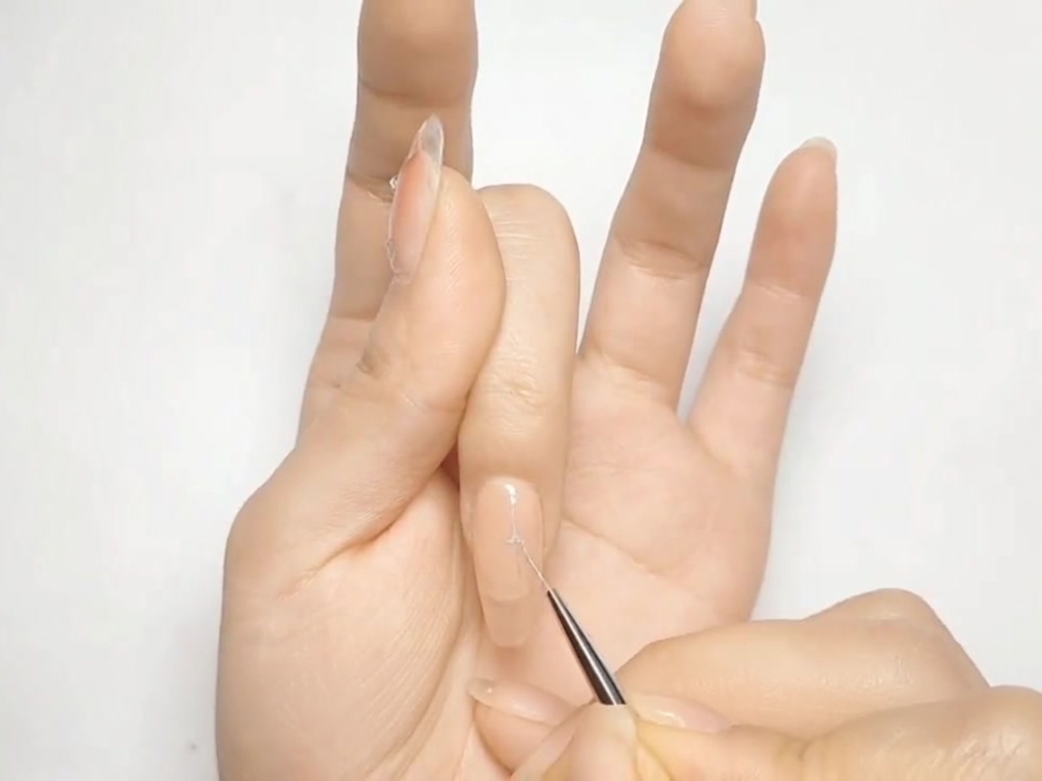 A thin nail art brush drawing a silver cross shape on a nude-painted nail.
